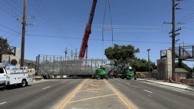 A crane positions the prefabricated steel pedestrian bridge over the roadway, demonstrating the efficiency of modular steel bridge construction.